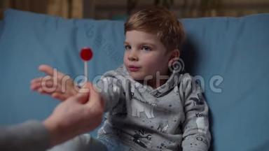 小男孩手拿糖果，晚上坐在舒适的沙发上，拿着棒棒糖到嘴边。 妈妈给我